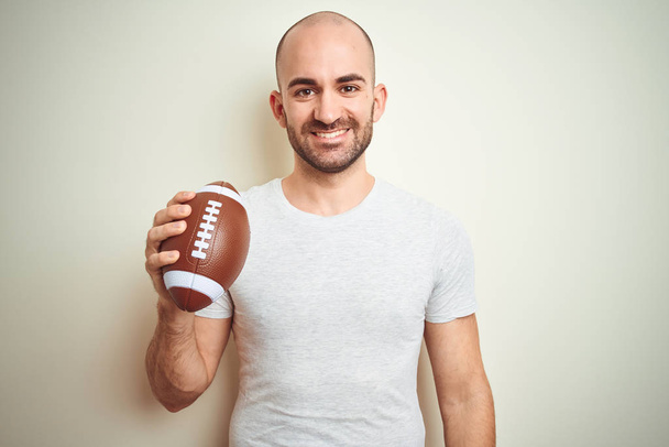 ラグビーアメリカンフットボールのボールを孤立した背景の上に持つ若者は、歯を見せる自信に満ちた笑顔で立ち上がり、笑顔を浮かべて立っている。 - 写真・画像