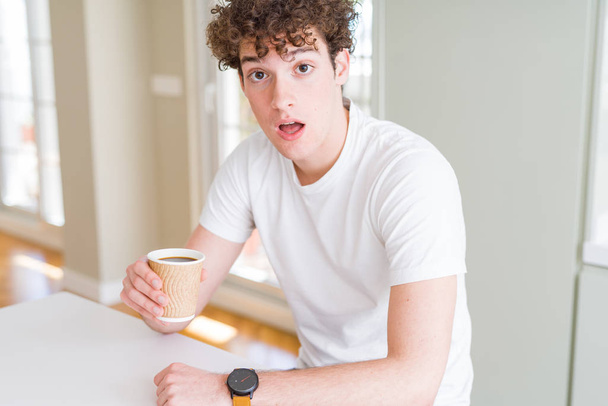 Jeune homme buvant emporter tasse de café à la maison effrayé dans le choc avec un visage surprise, effrayé et excité par l'expression de la peur
 - Photo, image
