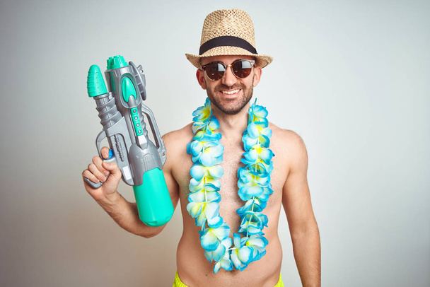 Young man wearing summer hat and hawaiian lei flowers holding water gun over isolated background with a happy face standing and smiling with a confident smile showing teeth - Photo, image
