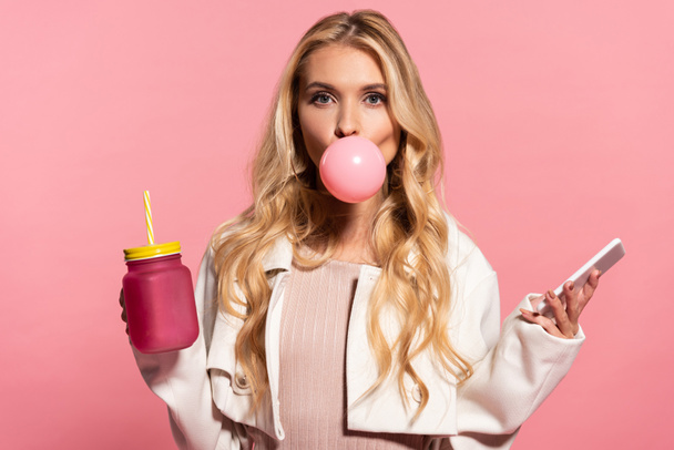 blonde young woman with pink bubble gum in mouth holding smartphone and jar isolated on pink - Photo, Image