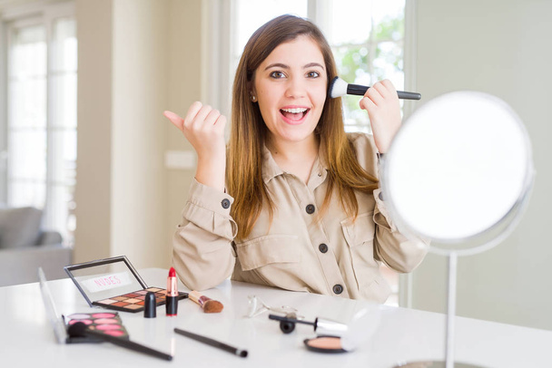 Mulher bonita usando maquiagem cosméticos aplicando cor usando escova apontando e mostrando com o polegar até o lado com o rosto feliz sorrindo
 - Foto, Imagem