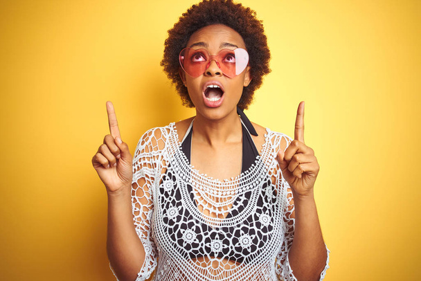 Bikini ve kalp şeklinde güneş gözlüğü giyen afro saçlı genç afro-amerikalı kadın şaşırttı ve baktı ve parmakları ile işaret ve kolları kaldırdı şaşırttı. - Fotoğraf, Görsel