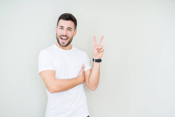 Young handsome man wearing casual white t-shirt over isolated background smiling with happy face winking at the camera doing victory sign. Number two. - Photo, Image