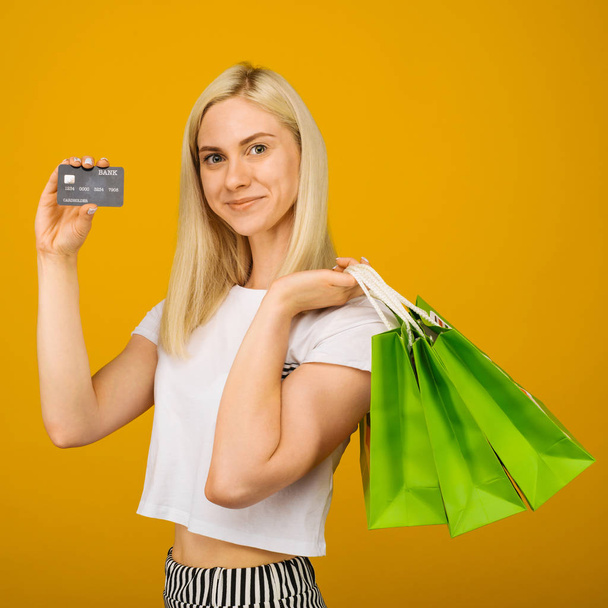 Közelről portré boldog fiatal gyönyörű szőke nő Holding hitelkártya és a zöld bevásárló táskák, nézett kamera, elszigetelt sárga háttér - Fotó, kép