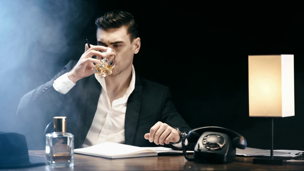 focused businessman writing in textbook and drinking whiskey while sitting at table on black - Imágenes, Vídeo