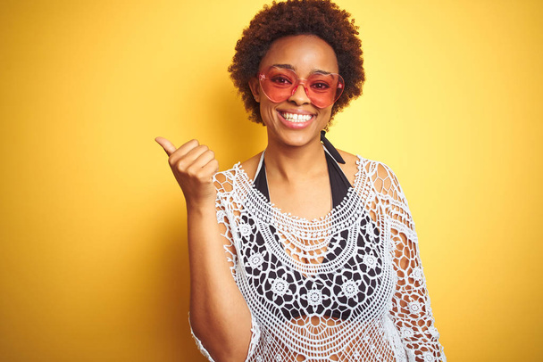 Fiatal afro-amerikai nő afro hajú bikini és szív alakú napszemüveg mosolyogva boldog arcát, és rámutatva, hogy az oldalsó hüvelykujjával fel. - Fotó, kép