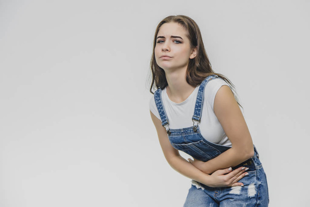 A tense young beautiful woman holds her arms on her stomach, feels painful emotions. She suffers from pain after hard work. A pretty white T-shirt, overalls, poses indoors. - Foto, imagen