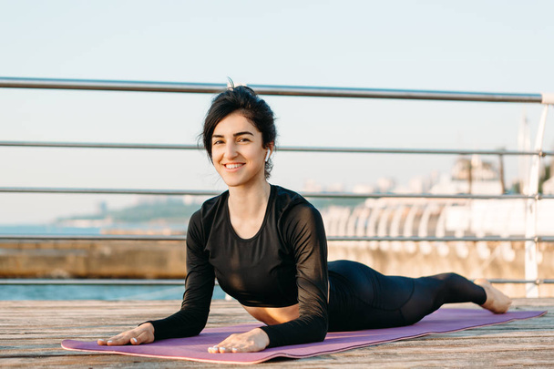 Beautiful young brunette woman in a black jumpsuit stretches on a pier at sunrise after exercising. - Photo, Image