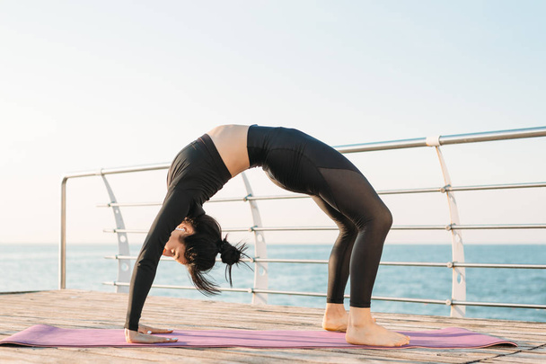 Belle jeune femme brune en combinaison noire pratiquant le yoga sur la plage au lever du soleil. Concept de bien-être et mode de vie sain
. - Photo, image
