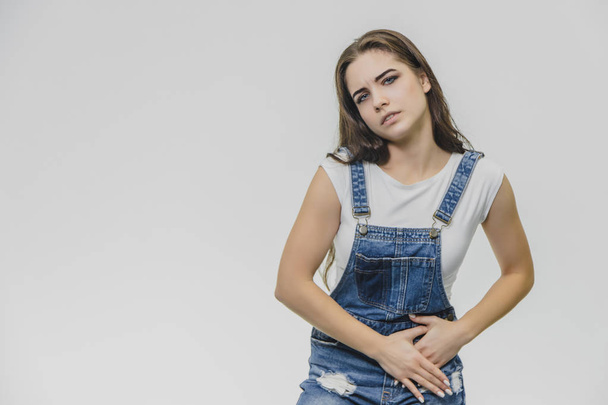 A tense young beautiful woman holds her arms on her stomach, feels painful emotions. She suffers from pain after hard work. A pretty white T-shirt, overalls, poses indoors. - Фото, изображение