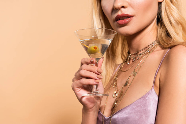 cropped view of elegant blonde woman in violet satin dress and necklace holding cocktail isolated on beige - Foto, afbeelding
