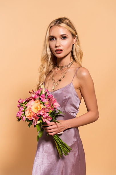 elegant blonde woman in violet satin dress and necklace holding bouquet of flowers isolated on beige - Foto, afbeelding