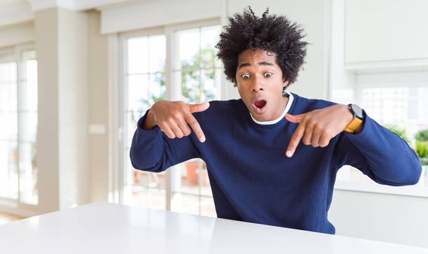 カジュアルなセーターを着た若いアフリカ系アメリカ人男性が、広告、驚いた顔、口を開いた指で指を下ろして下に座っている - 写真・画像