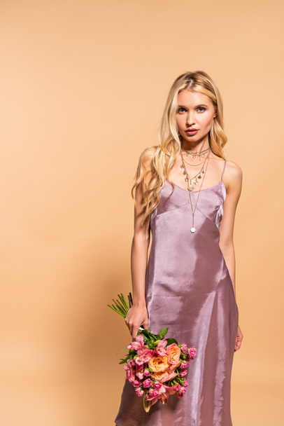 elegant woman in violet satin dress and necklace holding bouquet of flowers isolated on beige - Фото, зображення