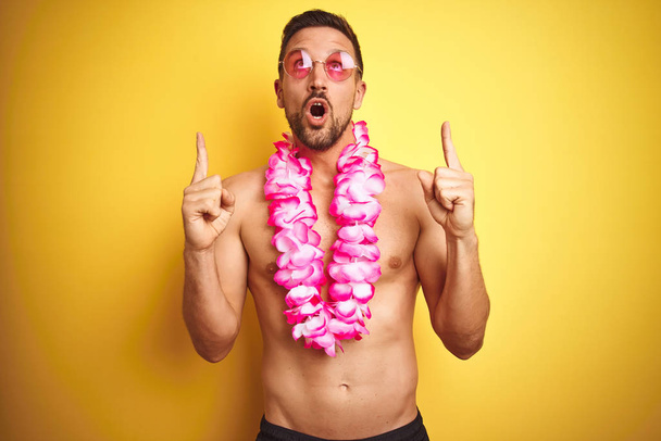 Молодой красивый мужчина без рубашки в солнечных очках и розовых гавайских леях на желтом фоне удивлен и удивлен, глядя вверх и указывая пальцами и поднятыми руками
. - Фото, изображение
