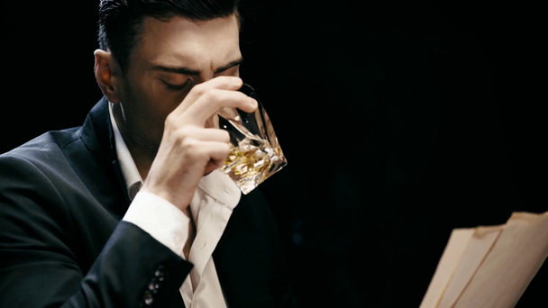 Konzentrierter Geschäftsmann liest Dokumente und trinkt Whiskey am Tisch mit Aschenbecher auf Schwarz - Filmmaterial, Video