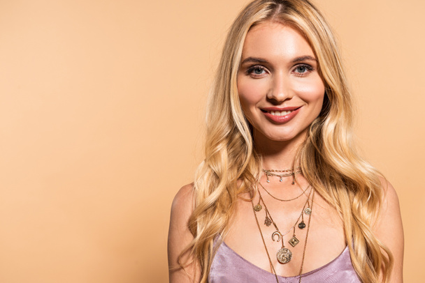 smiling blonde woman in violet satin dress and necklace isolated on beige - Foto, afbeelding