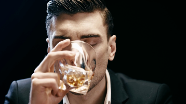 front view of serious businessman holding glass and drinking whiskey isolated on black - Video