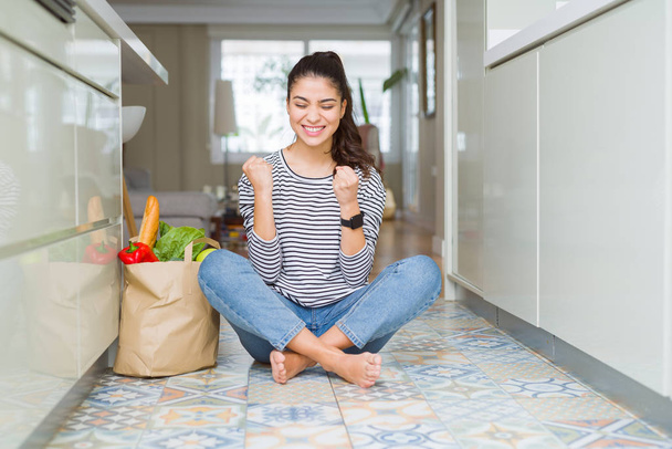 Young woman sitting on the kitchen floor with a paper bag full of fresh groceries very happy and excited doing winner gesture with arms raised, smiling and screaming for success. Celebration concept. - Foto, immagini
