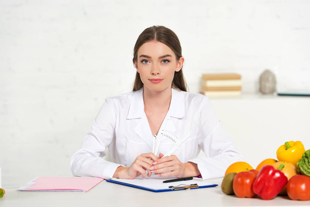 vista frontal del dietista en bata blanca en el lugar de trabajo con verduras, carpeta y portapapeles en la mesa
 - Foto, Imagen
