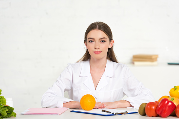 front view of dietitian in white coat at workplace with vegetables, folder and clipboard on table - Photo, Image