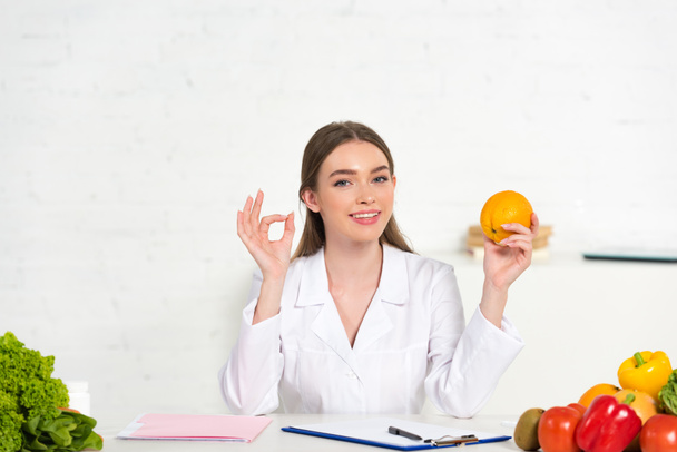 smiling dietitian in white coat holding orange and showing okay sign at workplace - Foto, Bild