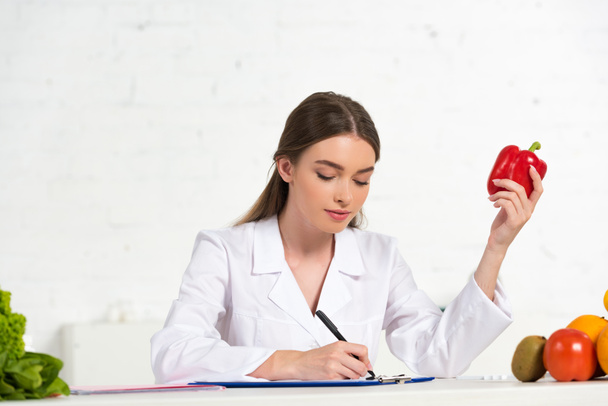 diététiste en manteau blanc tenant poivron rouge et écriture dans le presse-papiers sur le lieu de travail
 - Photo, image