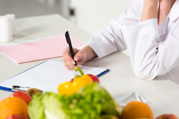 частичный вид диетолога в белом халате, пишущего в буфер обмена на рабочем месте с фруктами и овощами на столе
 - Фото, изображение