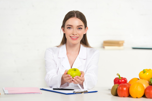front view of smiling dietitian in white coat holding apple at workplace - Fotoğraf, Görsel