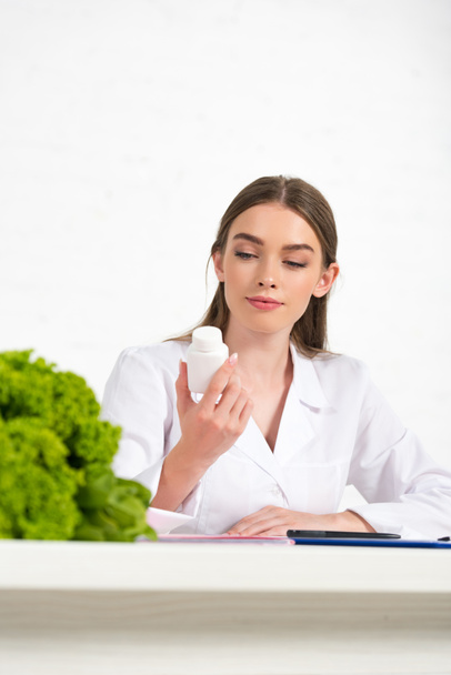 pensive dietitian in white coat holding pills at workplace - Photo, Image