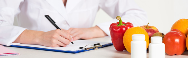 plano panorámico de dietista de capa blanca escrito en portapapeles en el lugar de trabajo con pastillas, frutas y verduras en la mesa
 - Foto, Imagen