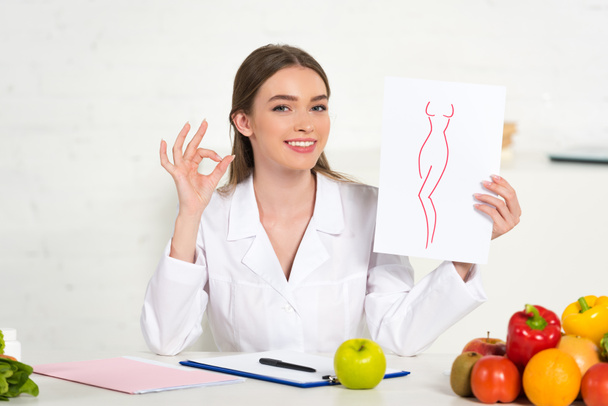 Lächelnder Diätassistent im weißen Mantel, der Papier mit Bild des perfekten Körpers hält und am Arbeitsplatz ein Okay-Schild mit Obst und Gemüse auf dem Tisch zeigt - Foto, Bild
