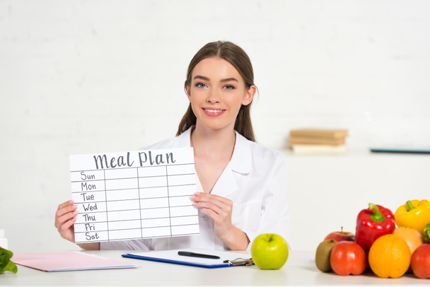 vista frontal del dietista sonriente en bata blanca que sostiene el plan de comidas en el lugar de trabajo con frutas y verduras en la mesa
 - Foto, imagen