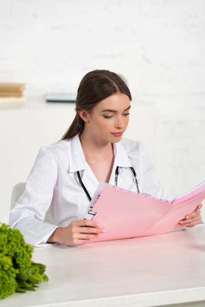 focused dietitian in white coat reading folder at workplace  - Photo, Image