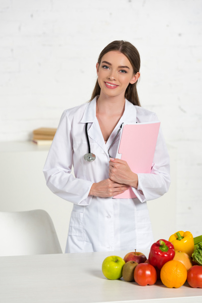 sorridente nutricionista atraente em casaco branco com equipamento segurando pasta perto da mesa com frutas e legumes frescos
 - Foto, Imagem