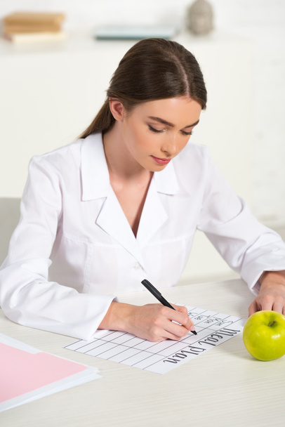 konzentrierte Ernährungsberaterin in weißem Mantel schreibt Speiseplan am Tisch mit grünem Apfel - Foto, Bild
