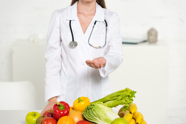 cropped view of dietitian in white coat with stethoscope standing with outstretched hand near fresh fruits and vegetables - Photo, Image