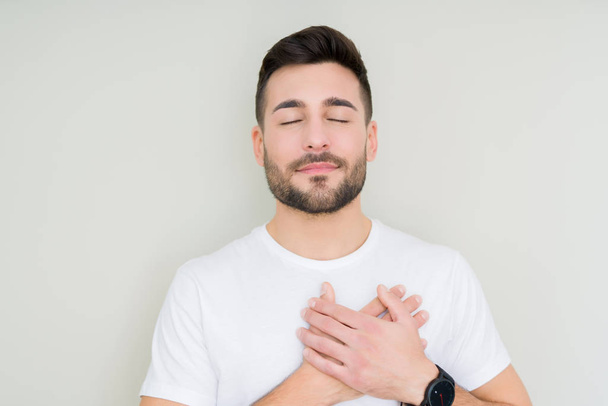 Young handsome man wearing casual white t-shirt over isolated background smiling with hands on chest with closed eyes and grateful gesture on face. Health concept. - Photo, Image