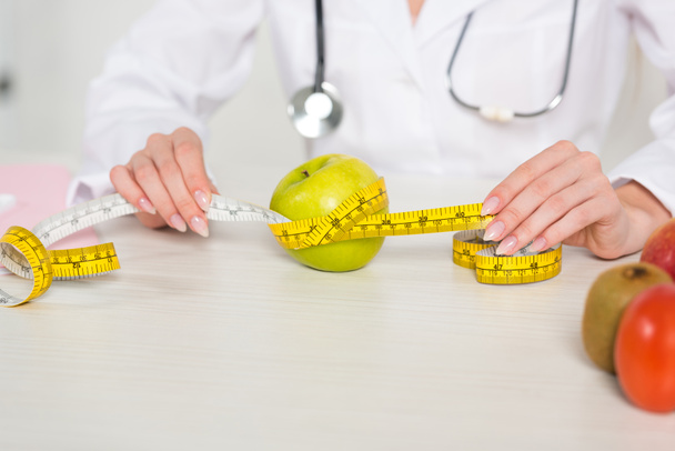vista cortada do nutricionista em fita métrica de retenção de casaco branco à mesa com frutas
 - Foto, Imagem