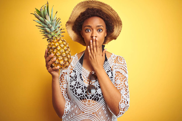 Joven mujer afroamericana con cabello afro sosteniendo piña sobre fondo amarillo aislado cubrir la boca con la mano conmocionada con vergüenza por error, expresión de miedo, asustado en silencio, concepto secreto
 - Foto, imagen