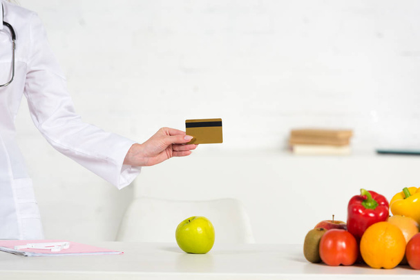 bijgesneden weergave van diëtist in witte vacht houden creditcard in de buurt van tafel met vers fruit en groenten - Foto, afbeelding
