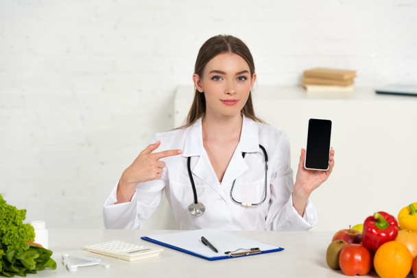 dietitian in white coat holding smartphone with blank screen and pointing with finger at workplace - Photo, Image