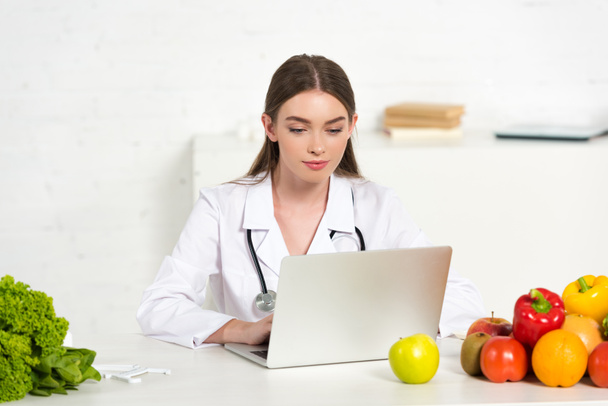 konzentrierte Ernährungsberaterin im weißen Kittel mit Laptop am Arbeitsplatz mit Obst und Gemüse - Foto, Bild