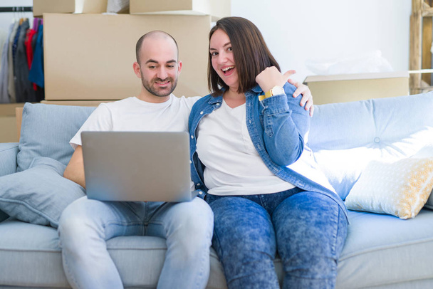 Young couple sitting on the sofa arround cardboard boxes moving to a new house using laptop pointing and showing with thumb up to the side with happy face smiling - Photo, Image