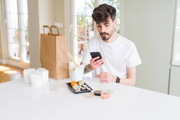 Mladý muž pojídáním asijských Suši z domácích dodávek a objednávání jídla pomocí smartphone aplikace se vystrašené hrůzou s překvapením, strachem a vzrušením výrazem strachu - Fotografie, Obrázek