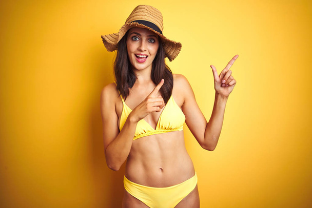 Mulher bonita usando biquíni amarelo e chapéu de verão sobre fundo amarelo isolado sorrindo e olhando para a câmera apontando com duas mãos e dedos para o lado
. - Foto, Imagem