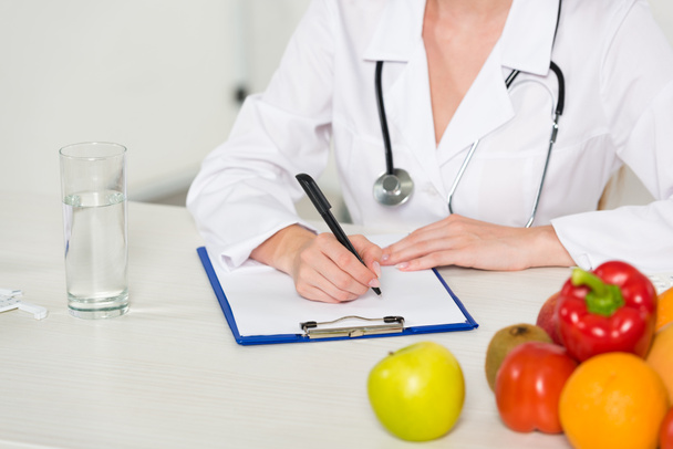 cropped view of dietitian in white coat writing in clipboard at workplace - Photo, image