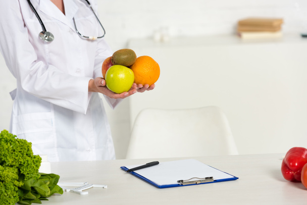 cropped view of dietitian in white coat holding fresh fruits at workplace - Foto, afbeelding