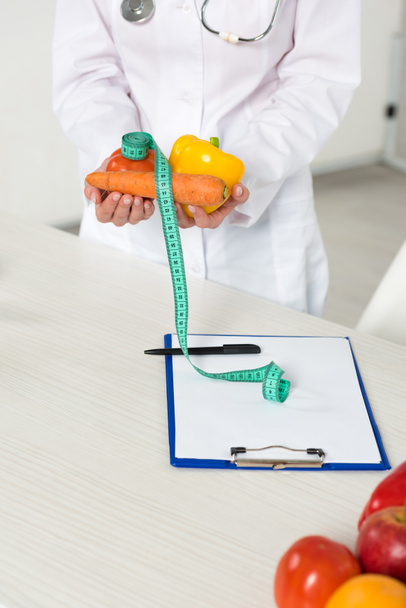 vue recadrée du diététiste en manteau blanc tenant des légumes et ruban à mesurer sur le lieu de travail
 - Photo, image