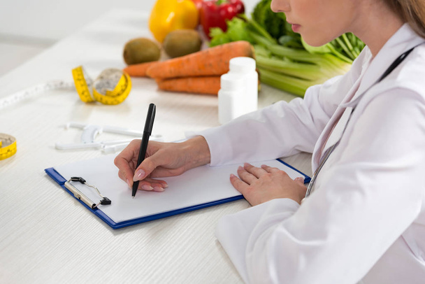 cropped view of dietitian in white coat writing in clipboard at workplace - Fotografie, Obrázek
